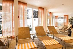 Heller Ruheraum mit Korbliegen mit orange gestreiften Kissen, großen Fenstern mit orangen Vorhängen und Blick auf verschneite Berge.