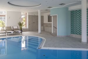 Moderner Innenpool im Hotel Upstalsboom mit blauem Wasser, gewölbter Decke, weißen Säulen, Liegestühlen und dekorativer Fliesenwand.