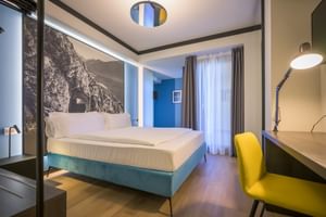 Modernes Hotelzimmer in der Villa Dirce Limone mit türkisfarbenem Bett, gelbem Stuhl, Schreibtischlampe, Berglandschafts-Wandbild und Balkonzugang.