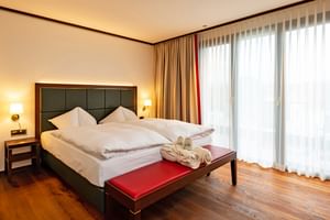 Modernes Doppelzimmer im Bad Horn Hotel mit dunklem Kopfteil, weißer Bettwäsche, roter Bank, Holzboden und großen Fenstern mit Vorhängen.