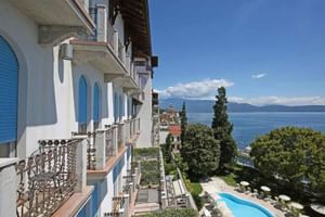 Hotel Savoy Gardone Terrasse mit blauen Fensterläden mit Blick auf den Gardasee, mit geschwungenem Pool umgeben von üppigen Gärten.