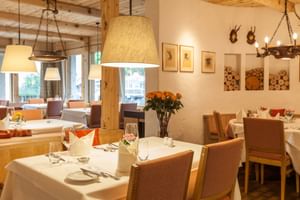 Elegant hotel restaurant with white-clothed tables, wooden chairs, pendant lamps, and alpine decor including antler mounts and stacked firewood.