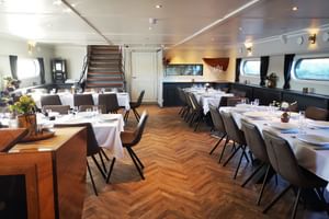 Elegantes Restaurant-Interieur auf der MS Magnifique IV mit weiß gedeckten Tischen, dunklen Stühlen, Parkettboden und Panoramafenstern.