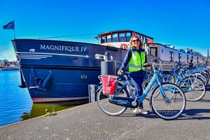 Frau in Warnweste mit Fahrrad steht neben dem Kreuzfahrtschiff MS Magnifique IV im Hafen unter blauem Himmel.