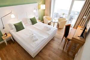 Modernes Hotel-Doppelzimmer mit weißem Bett, grünen Akzentkissen, Sitzbereich mit cremefarbenen Stühlen, Holzboden und Balkonzugang.