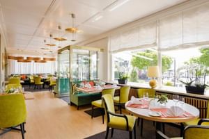 Modernes Restaurant mit runden Tischen, gelben Stühlen, grünen Sitzbänken und großen Fenstern zur Gartenterrasse.