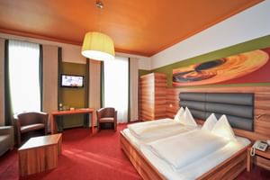 Modernes Doppelzimmer im Hotel Ertl mit Holzbett, rotem Teppich, oranger Decke, Sitzbereich und bunter Wandkunst.