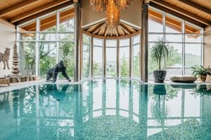 Eleganter Innenpool mit Holzdeckenbalken, großen Fenstern, Kristallleuchter und dekorativer Gorilla-Statue im Wellnessbereich.