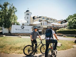 Zwei Radfahrer mit Fahrrädern vor dem Hotel Weisser Bär, einem weißen Gebäudekomplex mit markanten runden Turm und Außenterrasse.
