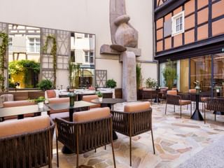 Außenterrasse im Hotel Le Colombier mit orangefarbenen Polsterstühlen, runden Tischen und dekorativer Steinskulptur umgeben von historischen Gebäuden.