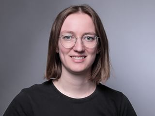 Porträt von Jael Loser mit schulterbraunem Haar, runder Brille und schwarzem Shirt, lächelnd in die Kamera vor grauem Hintergrund.