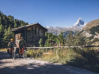 Deux cyclistes avec vélos sur une route de gravier près d'un chalet en bois, avec le Cervin emblématique visible en arrière-plan sous un ciel bleu clair.