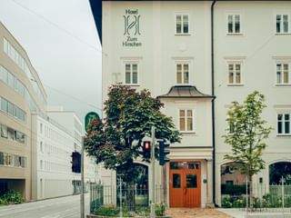 Weißes historisches Hotel zum Hirschen Gebäude in Salzburg mit verziertem Eingang, grünen Bäumen und modernen weißen Gebäuden links.