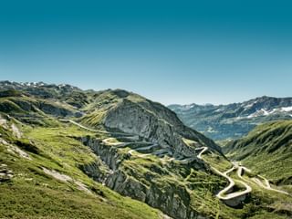 Vue aérienne de la route sinueuse du col du Gothard avec plusieurs virages en épingle traversant les montagnes alpines vertes sous un ciel bleu.