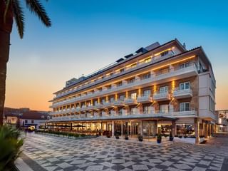 Modernes weißes Hotel Baia Gebäude mit mehreren Balkonen und warmer Beleuchtung bei Sonnenuntergang. Gemusterter Steinplatz im Vordergrund mit Palmen.