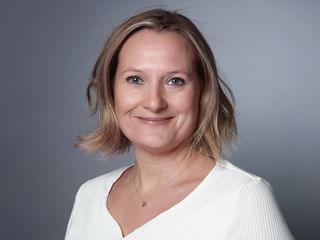 Portrait professionnel de Tanja Joss avec des cheveux blonds mi-longs, portant un haut blanc et un collier, souriant sur fond gris.
