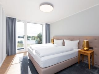 Helles Hotelzimmer mit großem Bett, grauen Vorhängen, Holznachttischen mit goldenen Lampen und Balkontür mit Blick auf die Müritz.