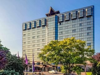Modernes Mercure Hotel in Koblenz mit Glasfassade und blauen Akzenten, umgeben von grünen Bäumen und Grünanlagen unter blauem Himmel.
