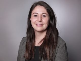 Professional portrait of Cindy Mayor Professional headshot of Cindy Mayor with long brown hair, wearing a gray blazer over a dark top, smiling at the camera against a gray background.