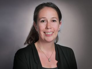 Professional portrait of Romaine Bayard Professional headshot of Romaine Bayard, a woman with brown hair wearing a dark blazer and necklace, smiling at the camera against a gray background.