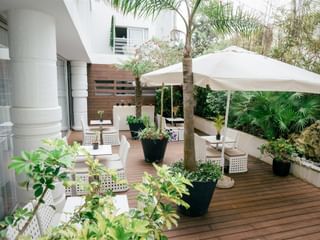 Holzterrasse im Macarico Beach Hotel mit weißen Korbmöbeln, großem Sonnenschirm, Topfpflanzen und tropischer Vegetation.
