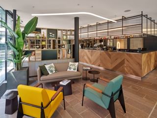 Zeitgenössische Hotelbar mit Holztheke, buntem Sitzbereich mit gelben und türkisfarbenen Stühlen, grauem Sofa und großer Pflanze.