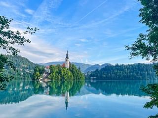 Lac de Bled en Slovénie avec petite île abritant une église au clocher blanc, entourée de collines boisées et montagnes sous ciel bleu.