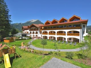 Großes Alpenhotel mit weißer Fassade und Holzbalkonen in grüner Anlage mit Spielplatz, umgeben von Bergen unter blauem Himmel.