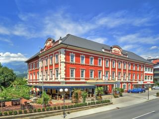 Hotel Ertl Spittal, ein rotes dreistöckiges Gebäude mit weißen Verzierungen und grauem Dach. Erdgeschoss-Terrasse mit Außenbestuhlung unter blauem Himmel.