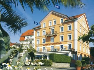 Gelbes Hotel Reutemann Gebäude in Lindau mit weißen Verzierungen, mehreren Stockwerken, Balkonen und Außenterrasse umgeben von Palmen und Blumen.