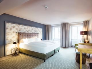 Moderne Hotel-Suite mit Doppelbett, grauem Polsterkopfteil, gemusterter Tapeten-Akzentwand, Holzschreibtisch und geometrischem Teppich.
