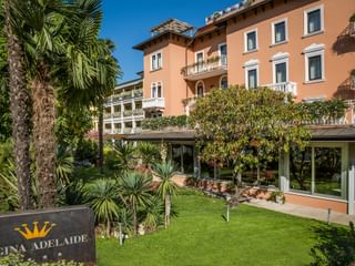 Elegantes korallfarbenes Hotel Regina Adelaide mit weißen Balkonen, umgeben von Palmen und gepflegten Gärten unter blauem Himmel.
