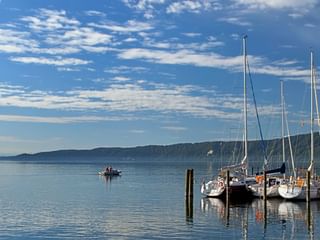 Panoramablick auf die Marina in Ludwigshafen am Bodensee mit Segelbooten am Holzsteg unter bewölktem blauen Himmel.