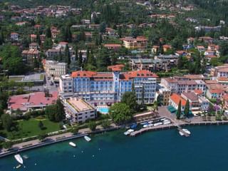 Luftaufnahme des Hotel Savoy Gardone, ein blau-oranges Hotel am Seeufer mit Pool, umgeben von mediterranen Gebäuden und Booten im Hafen.