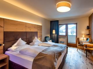 Modernes Hotelzimmer im Wiesnerhof Sterzing mit Holzkopfteil, weißer Bettwäsche, blauem Sessel, Schreibtisch und Bergblick-Fenster.