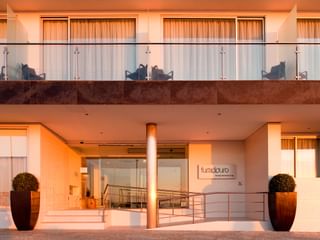Moderne Hotelfront mit Glaseingang, Balkonen mit Glasgeländern und dekorativen Pflanzgefäßen. Warme Beleuchtung erhellt die Fassade.