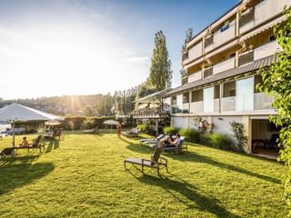 Mehrstöckiges Hotelgebäude mit Balkonen überblickt große grüne Rasenfläche mit Gartenmöbeln, Sonnenschirmen und entspannenden Gästen im goldenen Sonnenlicht.