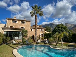 Hôtel Can Quatre en pierre à Sóller avec piscine extérieure, palmier et montagnes en arrière-plan sous ciel bleu et nuages blancs.
