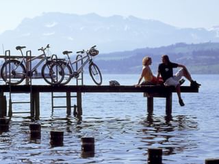 Zwei Radfahrer sitzen auf Holzsteg mit baumelnden Füßen über Seewasser. Drei Räder lehnen am Geländer mit Bergen im Hintergrund.