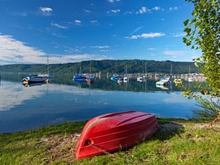Rotes umgedrehtes Boot am Grasesufer des Bodensees mit Segelbooten im ruhigen blauen Wasser und bewaldeten Hügeln im Hintergrund.