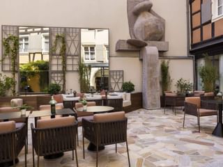 Cour d'hôtel moderne avec chaises en osier, tables rondes, sculpture en pierre et maisons à colombages traditionnelles à Colmar.