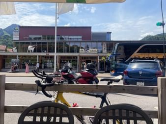 Blick vom Außensitzplatz am Bahnhof Gruyères mit Velo, parkenden Autos, modernem Bahnhofsgebäude und Bergen im Hintergrund.