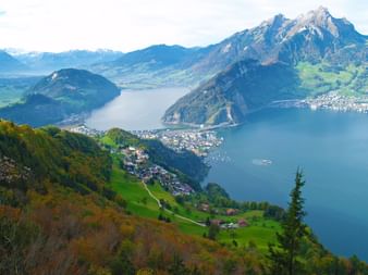 Vue aérienne du lac des Quatre-Cantons entouré de collines vertes et de montagnes enneigées. Des villages parsèment les rives.