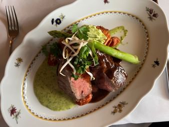 Geschnittenes Rindfleisch mit rosa Kern auf Blumenporzellan, garniert mit Spargel, Kräutern und grüner Sauce.