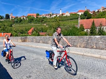 Zwei Radfahrer mit Tourenrädern und Packtaschen überqueren eine Kopfsteinpflasterbrücke im Taubertal mit mittelalterlicher Stadt.