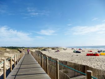 Holzsteg mit Holzzaun führt zu einem Sandstrand in Porto. Bunte Strandzelte und Menschen am Ufer unter blauem Himmel sichtbar.