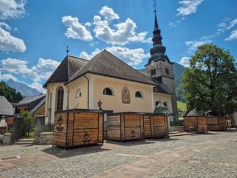 Gelbe Kirche mit dunklem Dach und hohem Glockenturm in Kranjska Gora. Holzmarktstände stehen davor auf einem Kopfsteinpflasterplatz unter blauem Himmel.