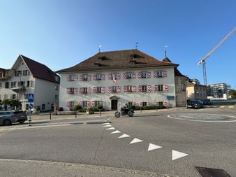 Historisches Gebäude in Saignelégier mit dekorativen bemalten Fensterläden mit regionalen Motiven. Ein Motorrad steht davor auf einem gepflasterten Platz.