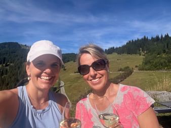 Zwei lächelnde Frauen mit Weingläsern im Freien auf der Gantrischhütte. Almwiesen und bewaldete Hügel unter blauem Himmel im Hintergrund.