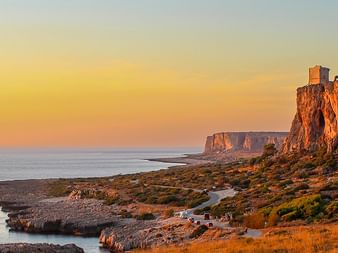 Golden sunset over dramatic coastal cliffs in Sicily with ancient stone buildings perched on rocky outcrops overlooking the Mediterranean Sea.